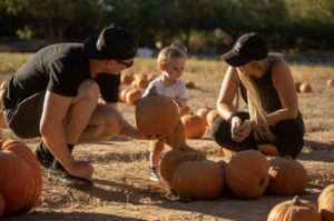Two adults crouching in the pumpkin patch hand a toddler a pumpkin.