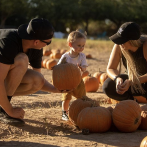Two adults crouching in the pumpkin patch hand a toddler a pumpkin.